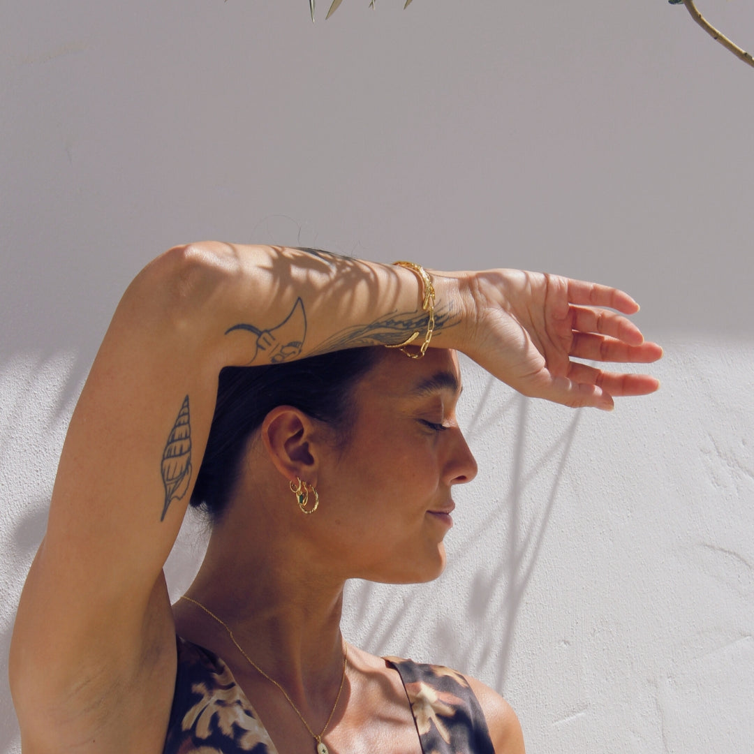 Woman with tattoos and gold jewelry against a neutral outdoor background