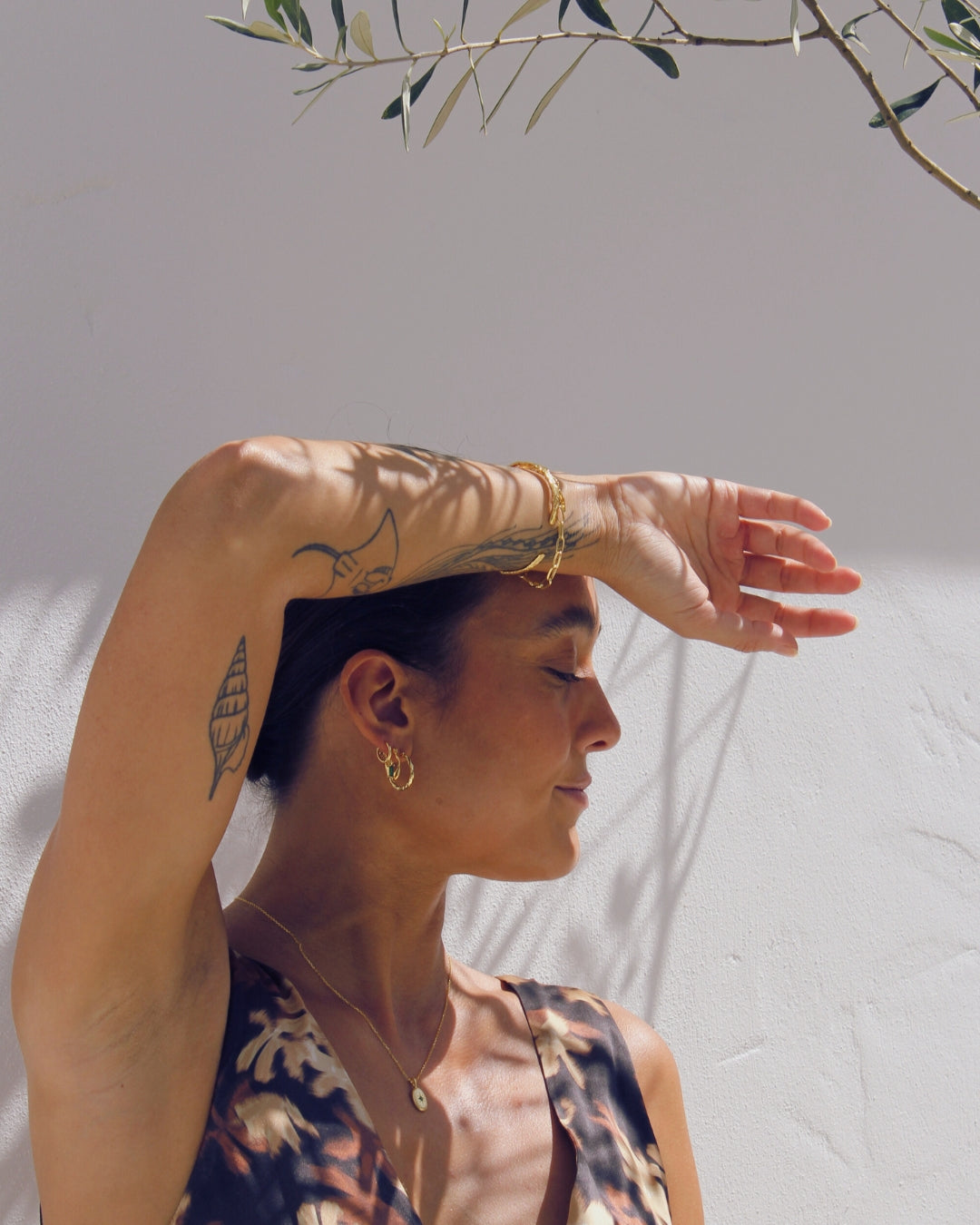 Woman with tattoos and gold jewelry against a neutral outdoor background