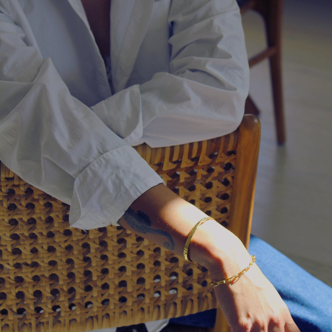 Person wearing a white shirt and blue jeans sitting on a wicker chair with two bracelets one of which is the Avalon Cuff