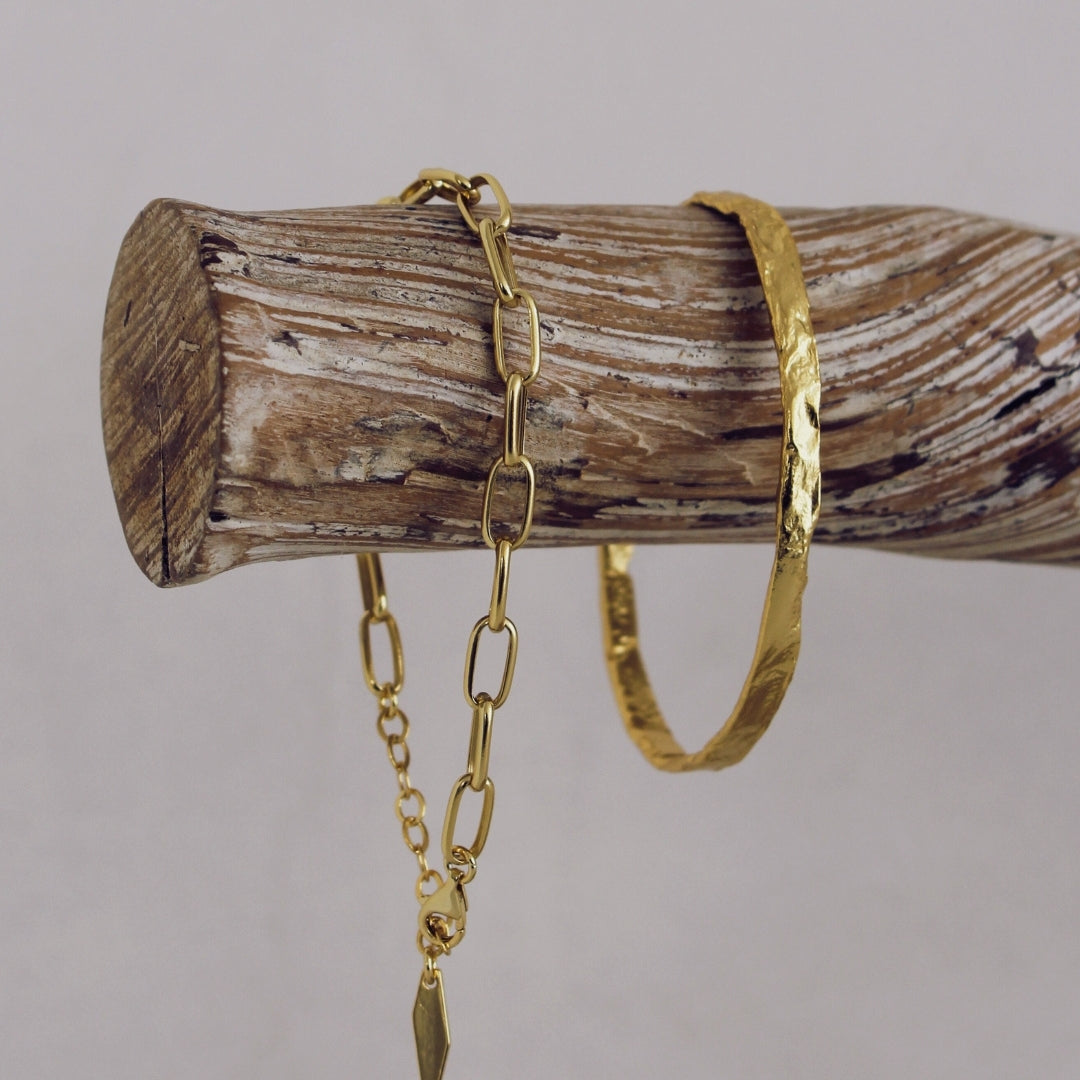 Gold chain bracelet and Avalon Cuff on a wooden log against a grey background