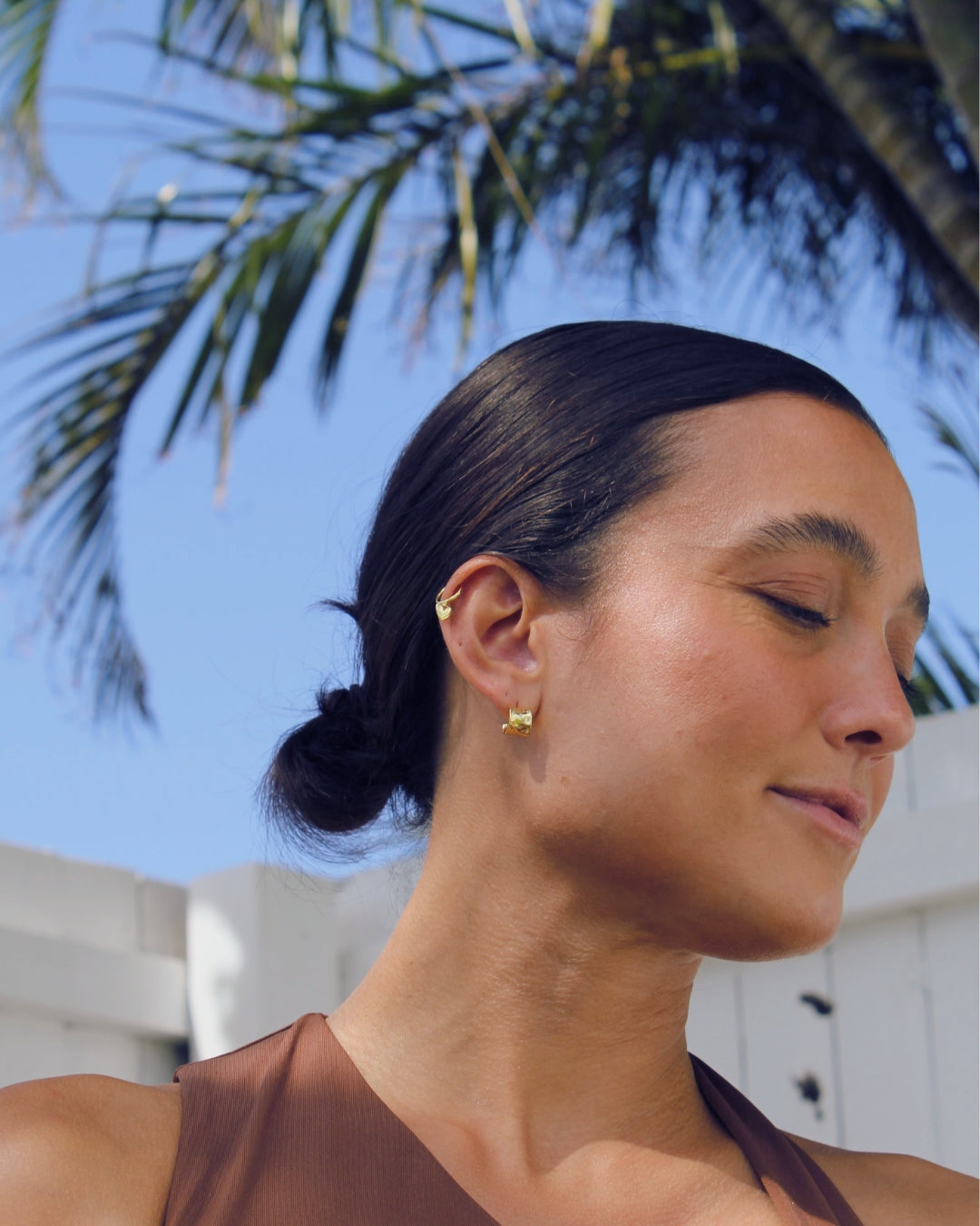 Woman with a bun wearing gold earrings against a palm tree background