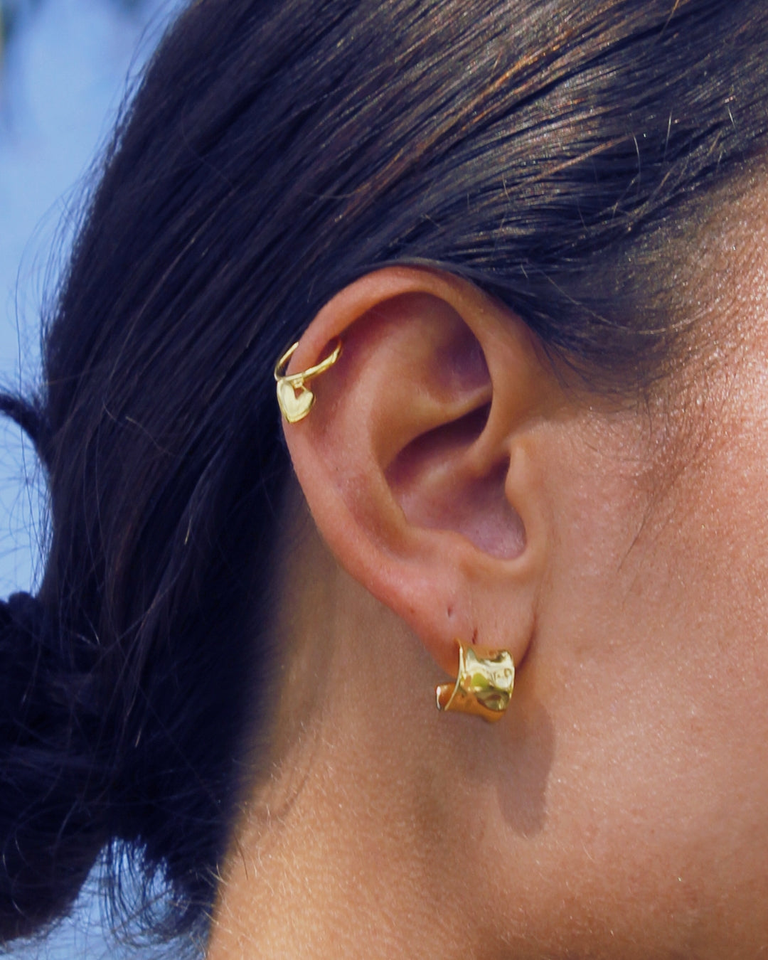 Close-up of an ear wearing gold pendant heart hoop earrings with a blurred background