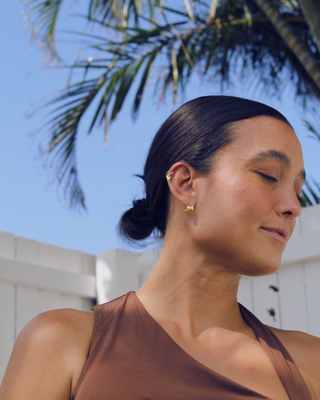 Woman with a bun hairstyle and 18k gold earrings against a palm tree and blue sky background