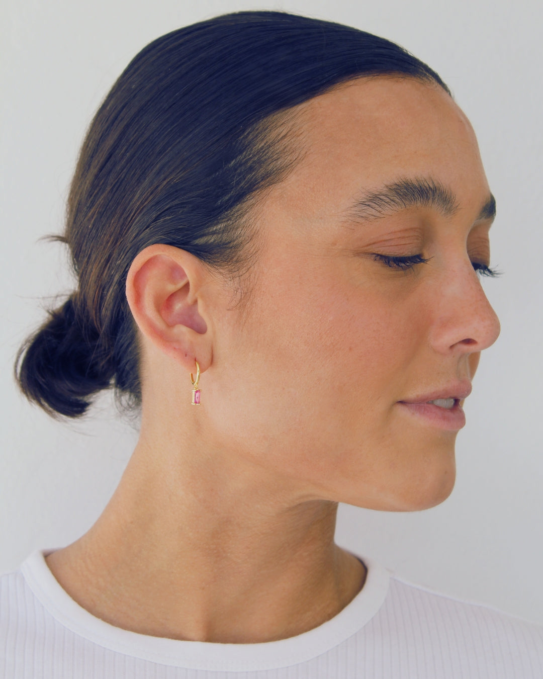 Woman with bun wearing a gold pendant hoop earring with a pink gemstone against a white background