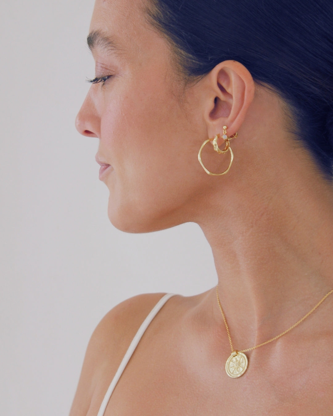 Woman wearing gold hoop earrings and a necklace with a coin pendant against a plain background