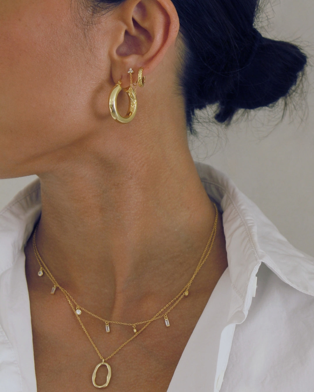 Close-up of a person wearing gold hoop earrings and necklaces against a neutral background