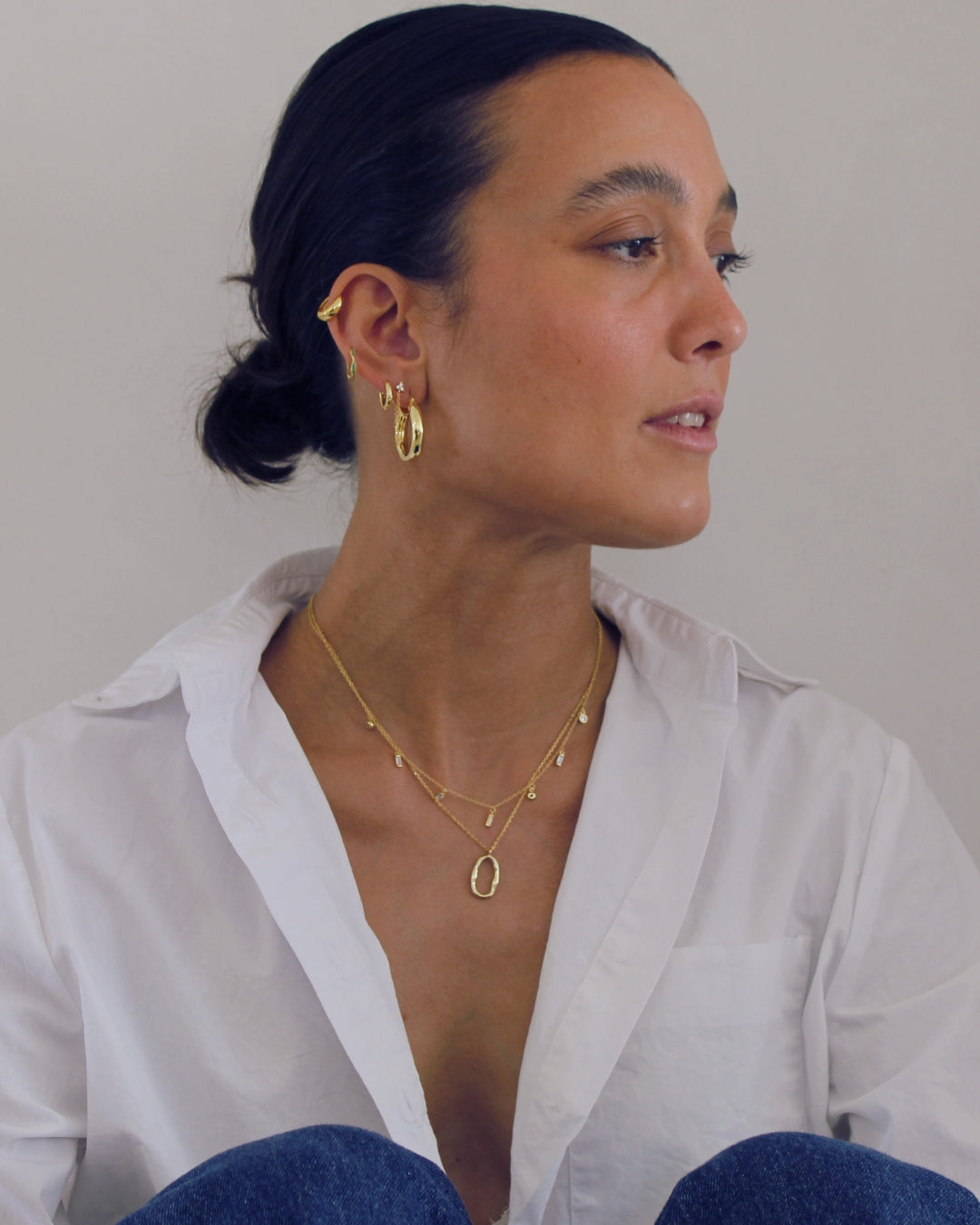 Woman wearing gold jewelry including earrings and a necklace sitting against a wall.