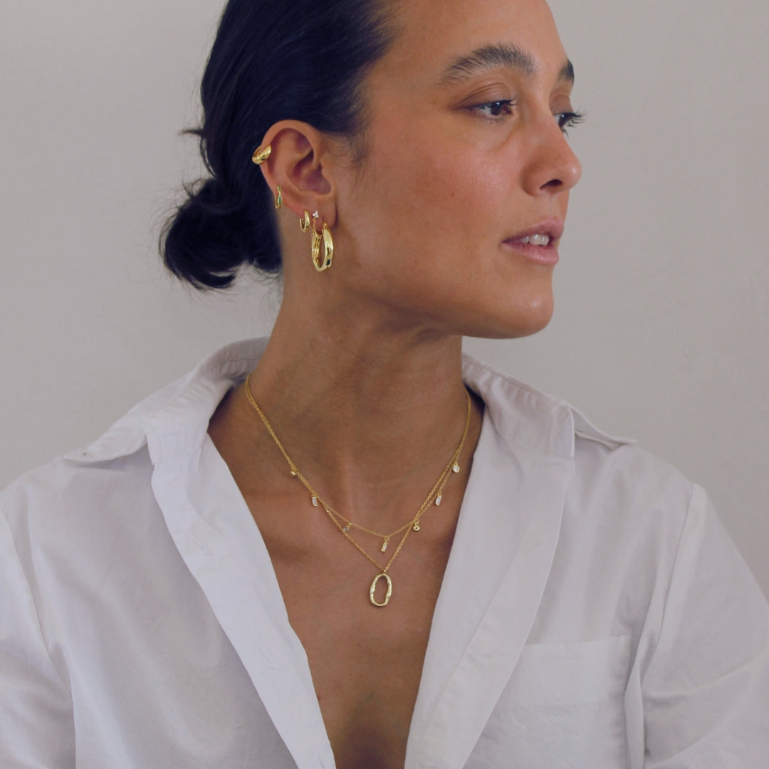 Woman wearing gold jewelry including earrings and a necklace, sitting against a plain background.