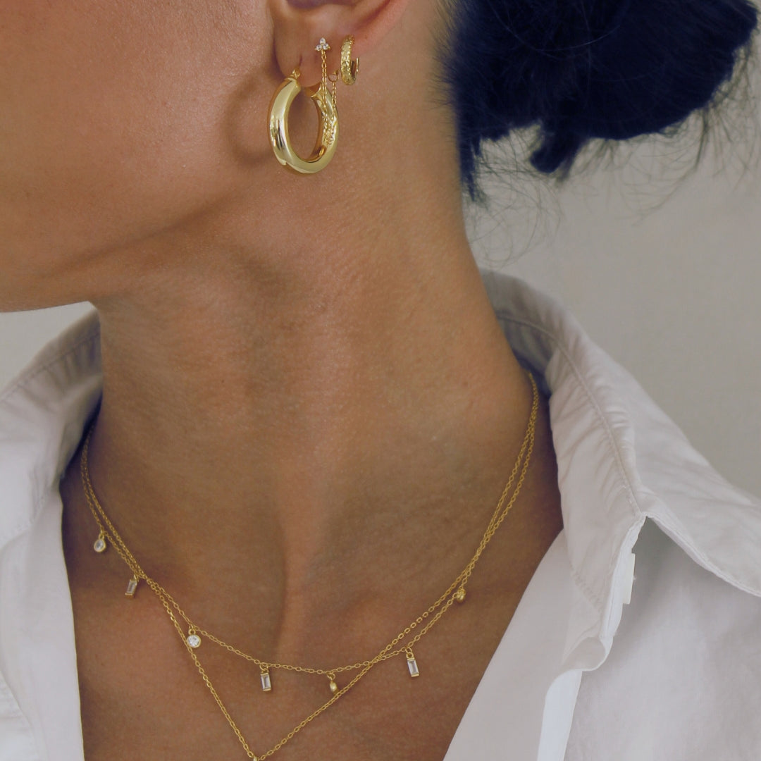 Close-up of a person wearing gold hoop earrings and necklaces including the Mae against a neutral background
