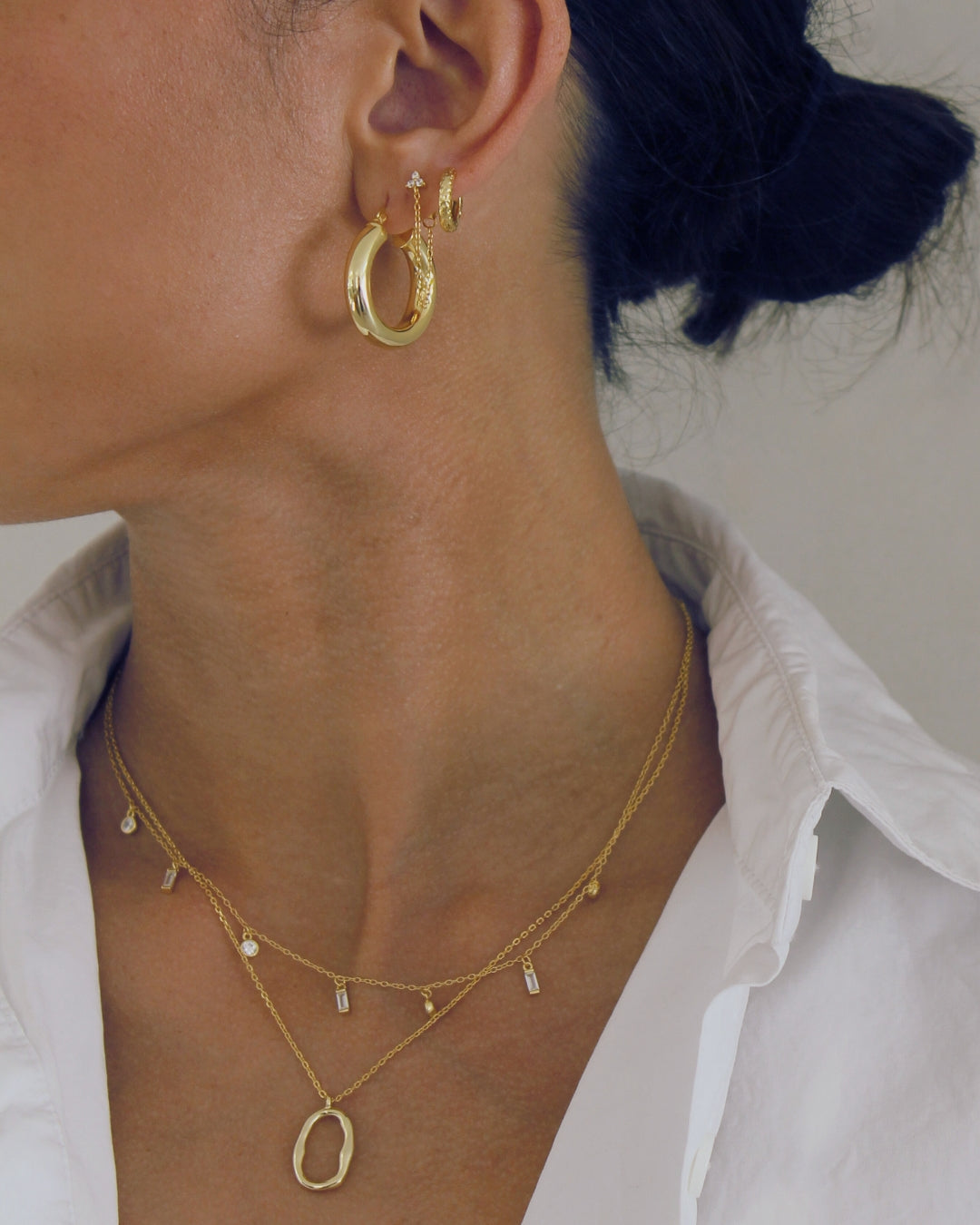 Close-up of a person wearing gold hoop earrings and necklaces including the Mae against a neutral background