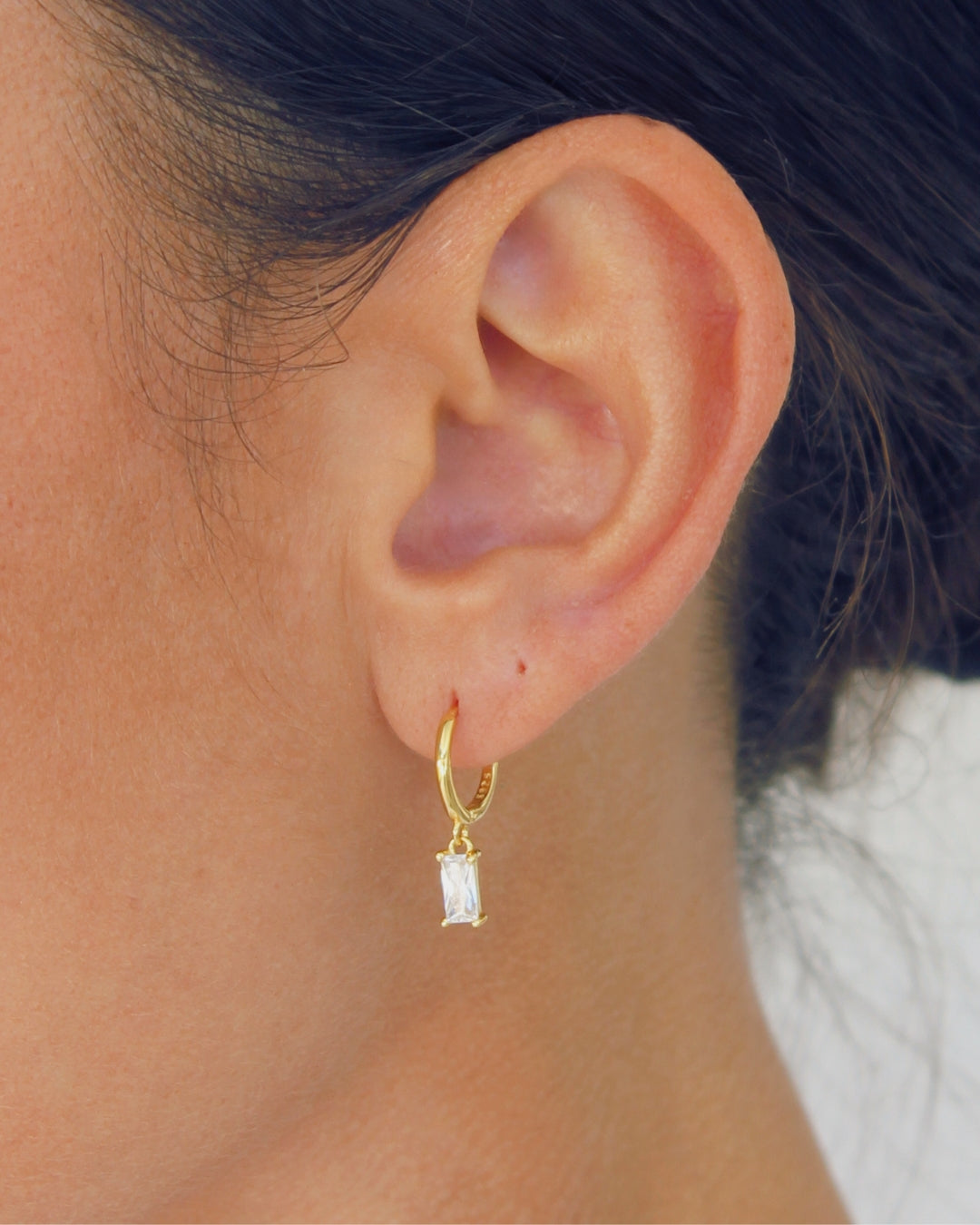 Close up of a Gold hoop earring with a clear zirconia pendant worn by a person.
