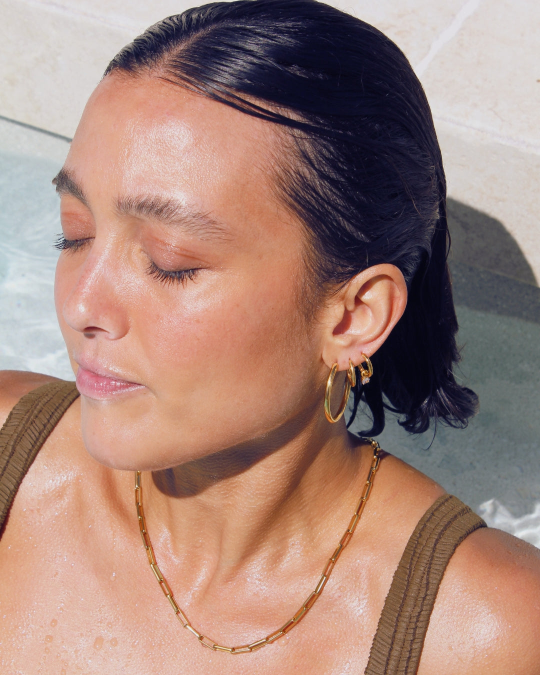 Woman with closed eyes, wearing a gold pendant hoop earring and necklace, sitting in a pool.