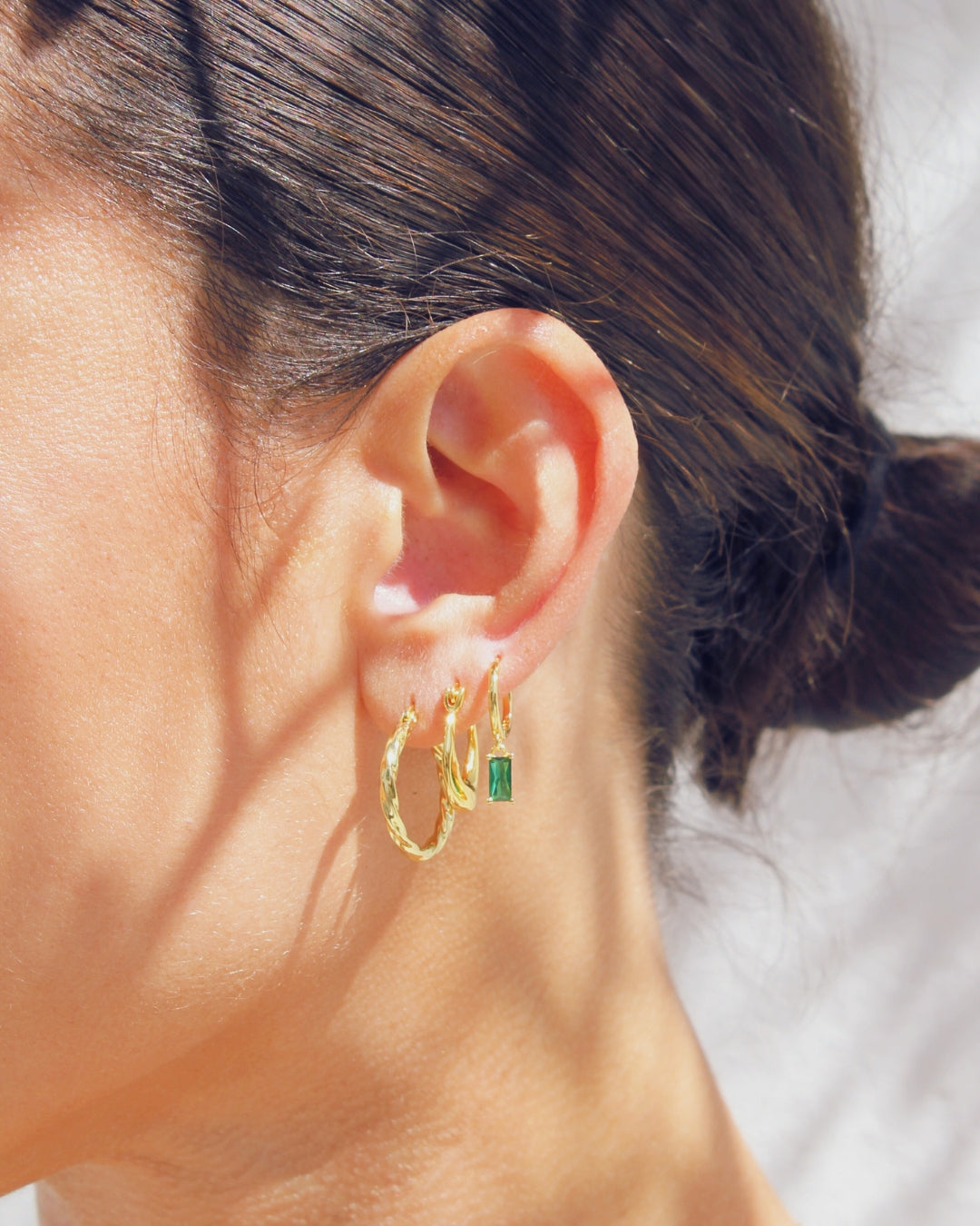 Close-up of an ear wearing gold pendant hoop earrings with a green gemstone.