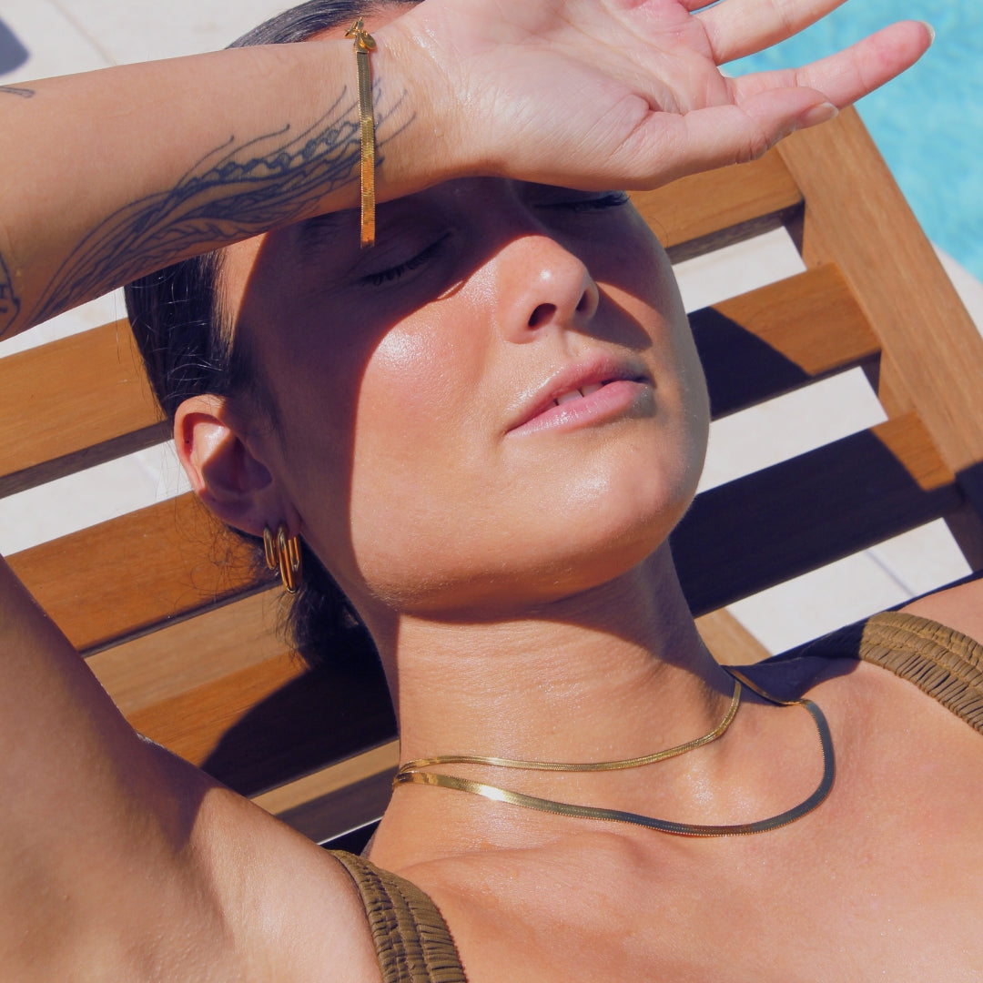 Woman shielding her eyes from the sun with a hand, wearing 18 k water safe gold jewellery, on a pool side recliner