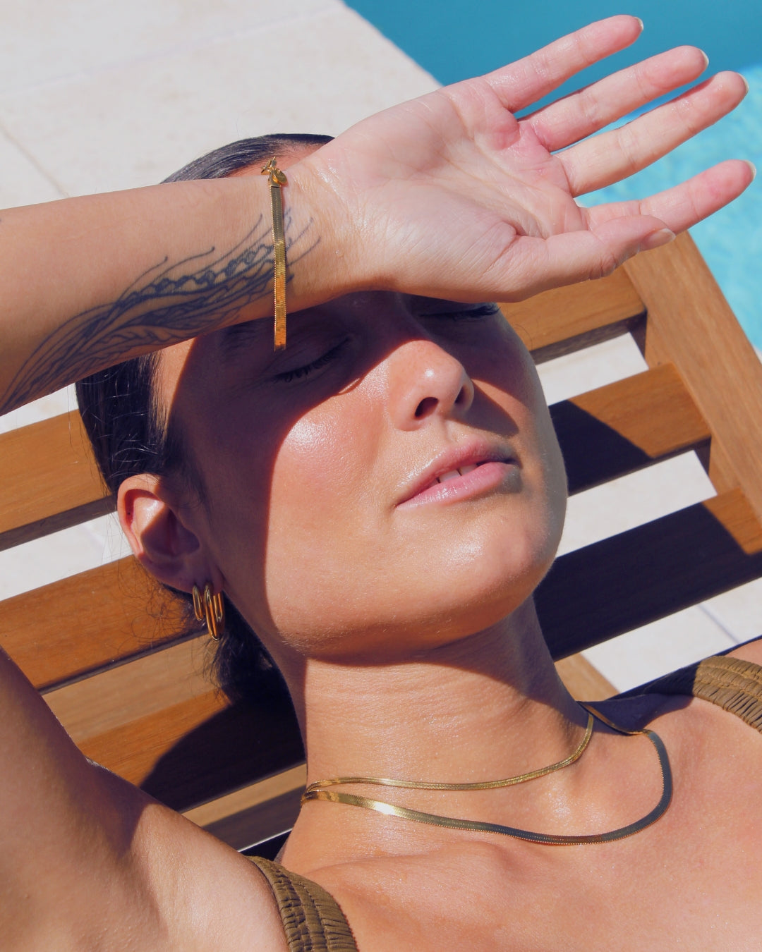 Woman shielding her eyes from the sun with a hand, laying by a pool with 18k waterproof jewellery.