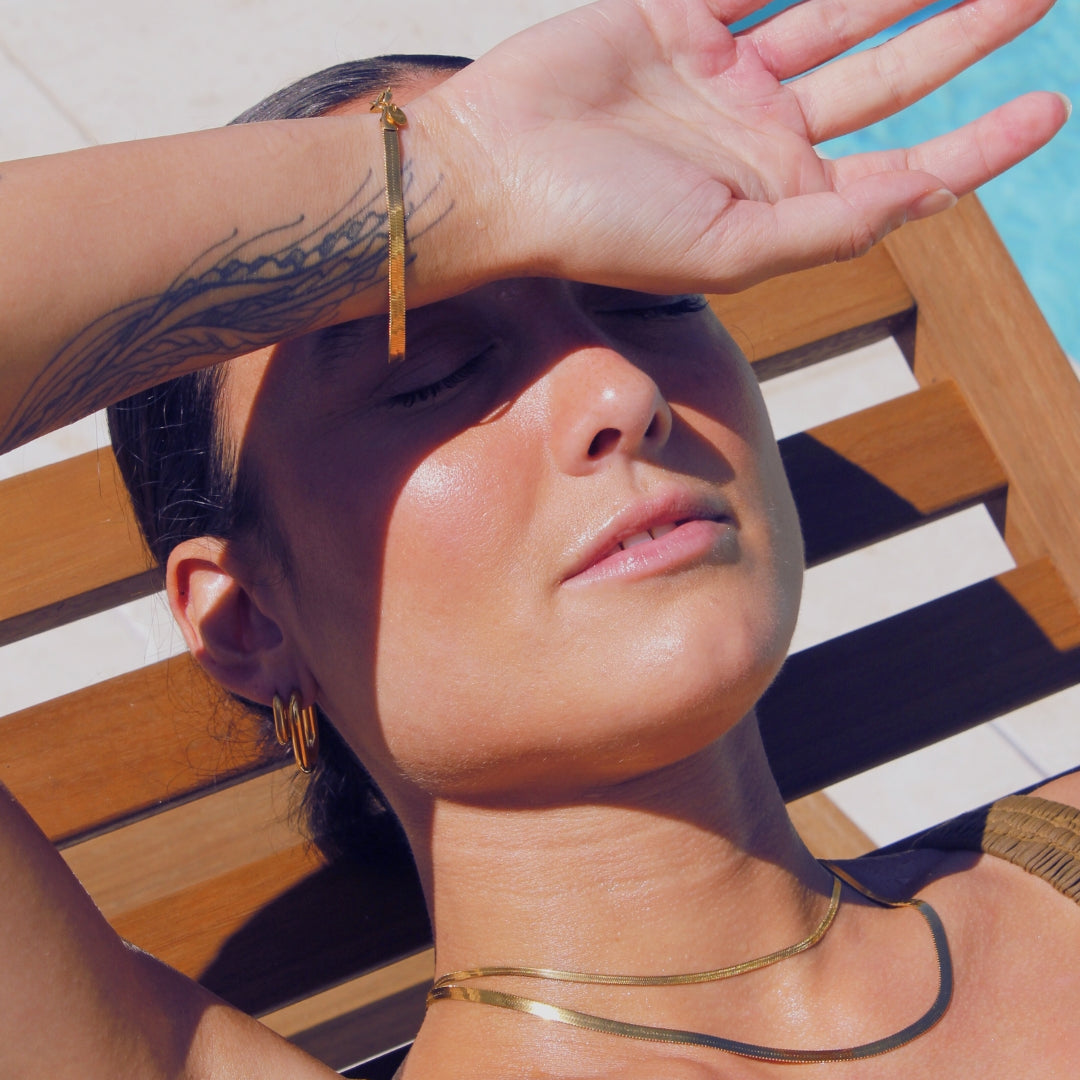 Woman shielding her eyes from the sun with a hand, sitting on a striped chair by a pool wearing 18k gold waterproof jewellery.