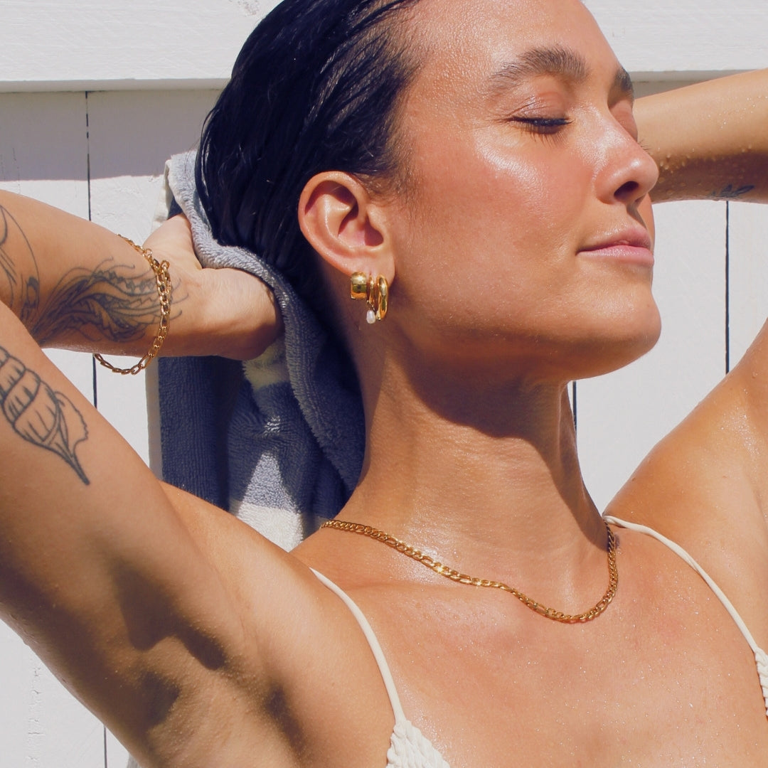 Woman with tattoos and waterproof gold jewellery against a white wooden background