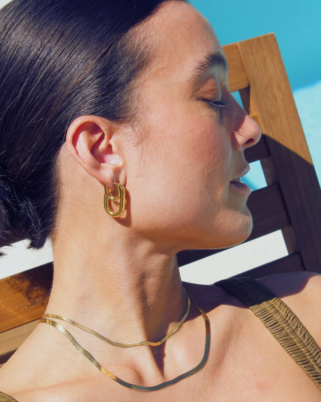 Woman wearing gold hoop earrings and necklace laying next to a pool on a recliner