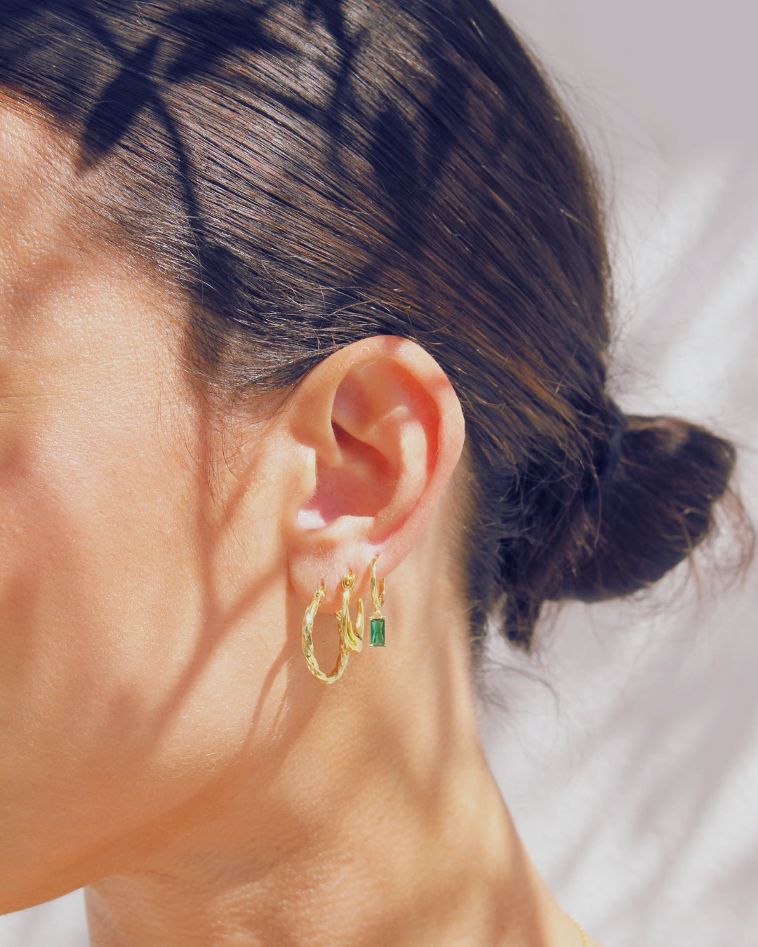 Close-up of an ear wearing gold hoop earrings with a green gemstone on a blurred background