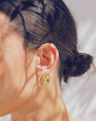 Close-up of an ear wearing gold hoop earrings with a green gemstone on a blurred background