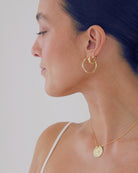 Woman wearing gold hoop earrings and a necklace with a coin pendant against a plain background