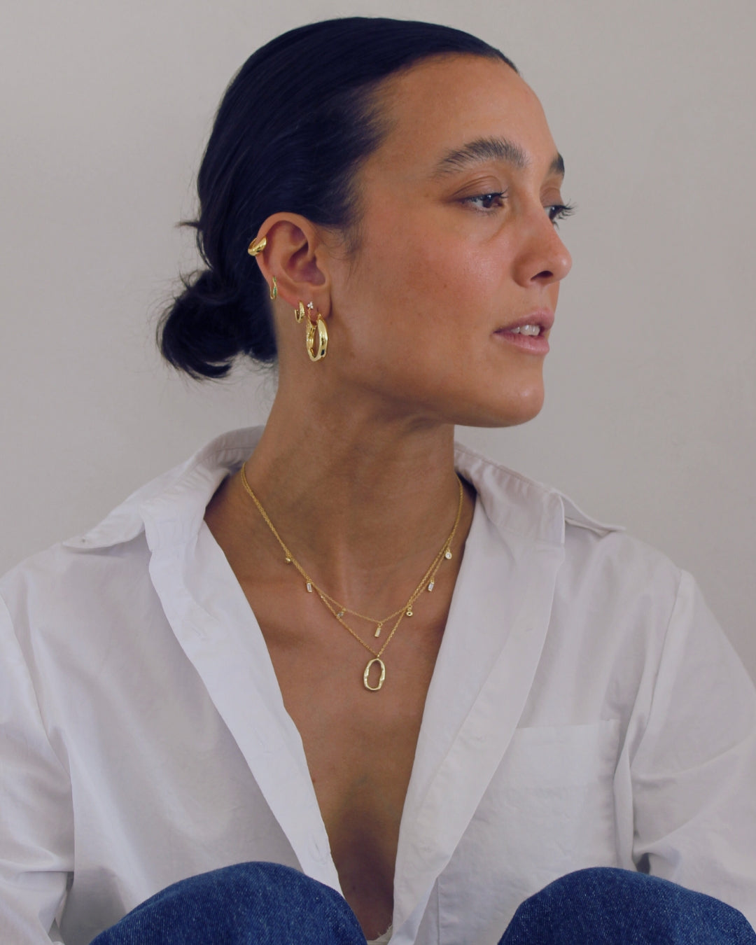Woman wearing gold jewelry including earrings and a necklace, sitting against a plain background.
