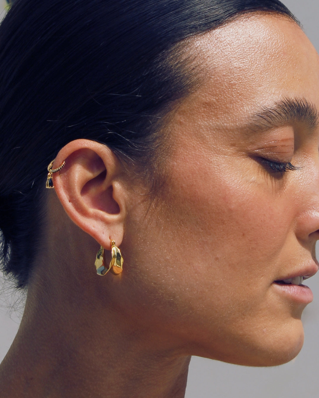 Close-up of a woman wearing  the Stella 18k gold hoop earrings with a neutral background