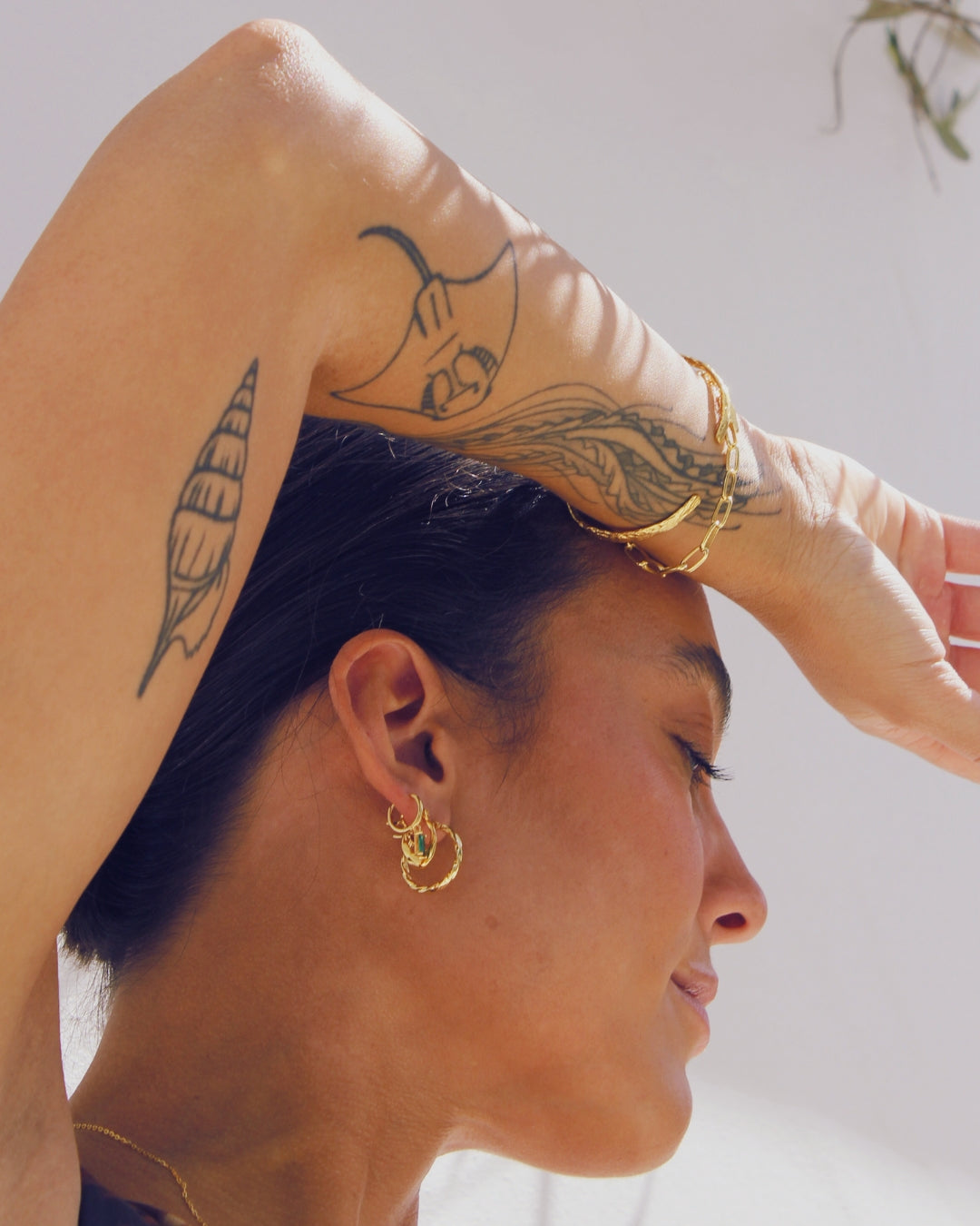 Woman with tattoos on her arm wearing gold jewelry, including earrings and a bracelet.