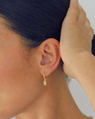 Close-up of an ear wearing a gold earring with a clear gemstone against a neutral background