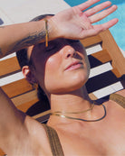 Woman shielding her eyes from the sun with a hand, wearing 18 k water safe gold jewellery, on a pool side recliner