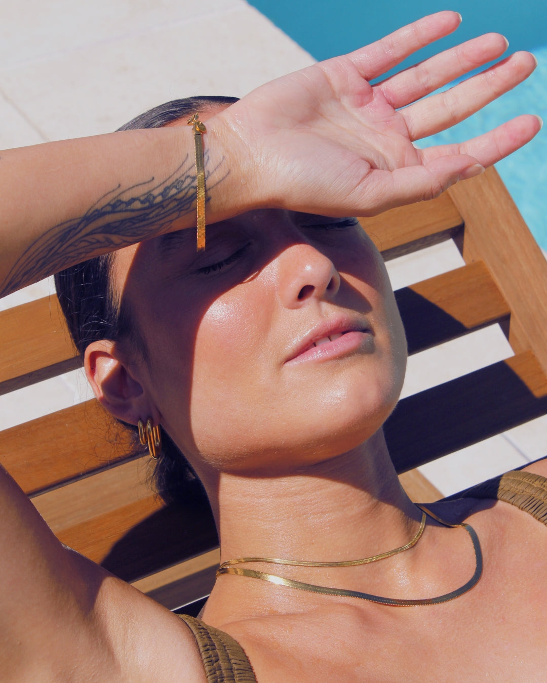Woman shielding her eyes from the sun with a hand, sitting on a striped chair by a pool wearing 18k gold waterproof jewellery.