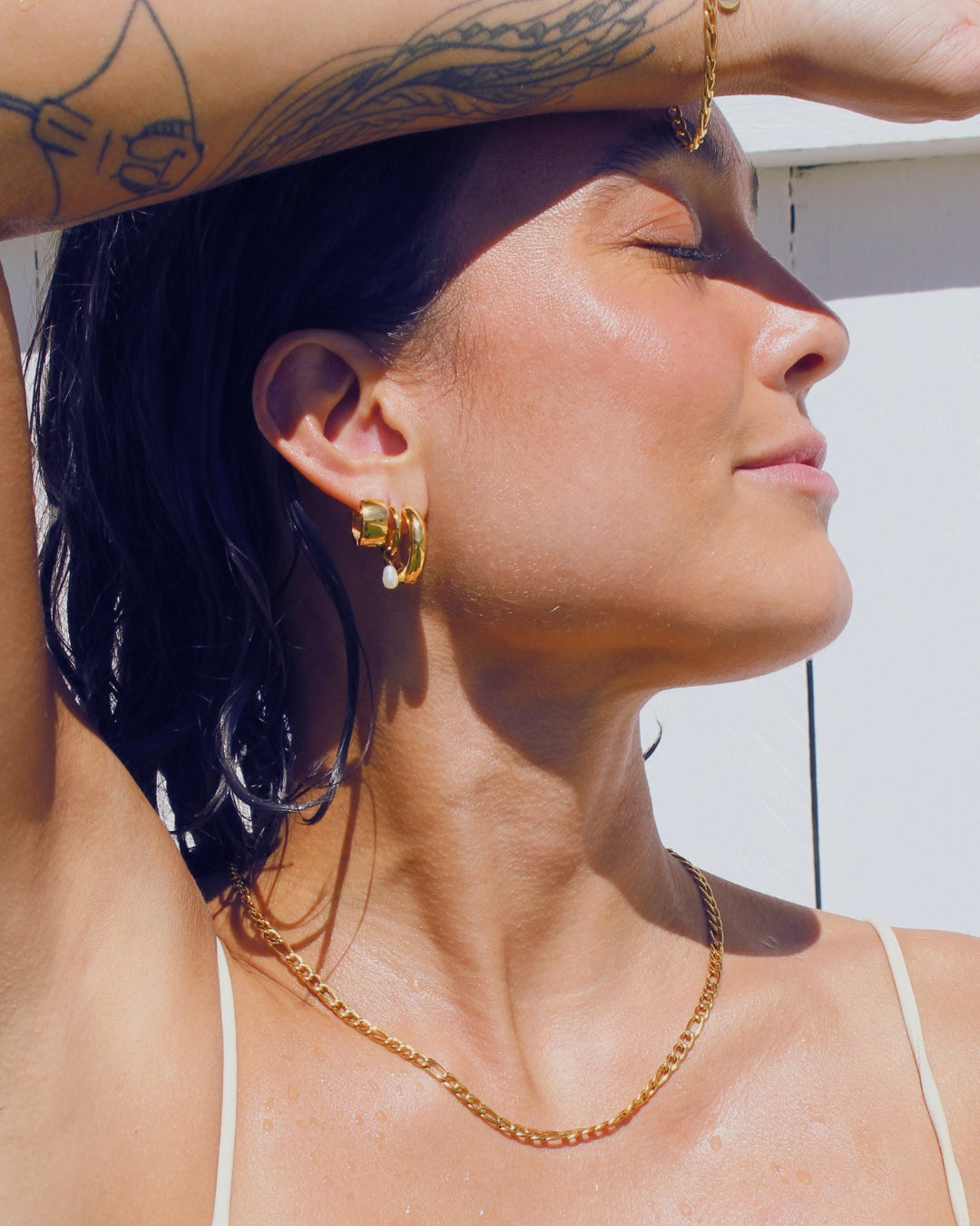 Woman with tattoos shielding her eyes from the sun, wearing gold waterproof jewellery.
