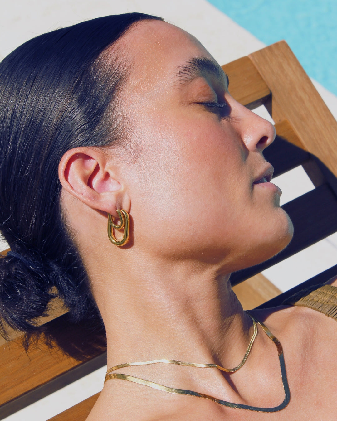 Woman wearing gold hoop earrings and a gold necklace, laying next to a pool